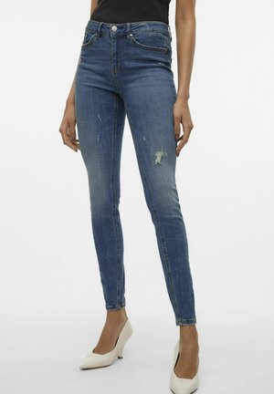 Skinny blue denim jeans with a mid-rise waist, featuring distressed details and classic five-pocket design. Paired with white heels.