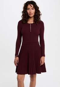Vestido burdeos de canalé con mangas largas, corsé ajustado, detalle de botones y falda acampanada con tablas. Tejido suave con una textura lisa.