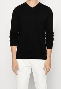 Černý svetr s dlouhými rukávy a výstřihem do V, vyrobený z měkké látky, s žebrovanými manžetami a spodním lemem, nošený přes bílé kalhoty, ukazující přiléhavý střih.