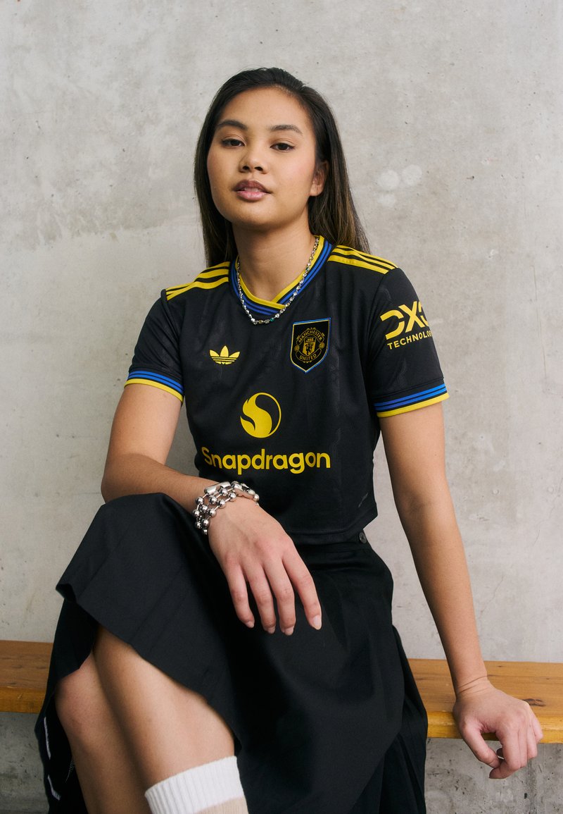 Camisola preta de manga curta com detalhes em azul e amarelo, apresentando o logótipo "Snapdragon" e o emblema da Adidas. Combinando com uma saia preta fluida.