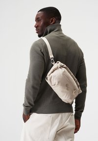 Beige Umhängetasche aus weichem Material, mit einer Reißverschlusstasche und zwei dekorativen Knöpfen. Getragen über einem grauen Pullover.