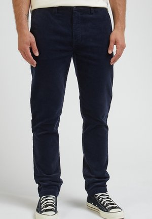 Pantalon en velours côtelé bleu marine avec une coupe slim, doté d'un devant plat, de passants de ceinture et d'une fermeture éclair, associé à des baskets noires basses.