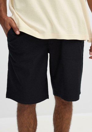 Hombre vistiendo pantalones cortos negros hasta la rodilla y una camisa color crema, con una mano en el bolsillo de los pantalones cortos, sobre un fondo liso.