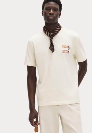 Hombre con camiseta crema con texto y gráfico "LIBRE IBIZA", pantalones crema y un pañuelo marrón a rayas en el cuello, sosteniendo un accesorio pequeño de cuero.