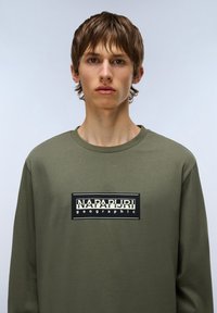 Maglietta a maniche lunghe color verde oliva in cotone, con un logo rettangolare bianco e nero sul petto con design testuale.