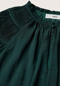 Blouse vert foncé avec détails froncés aux épaules, encolure ronde avec tissu froncé et fermeture à bouton unique recouvert de tissu à l'avant.