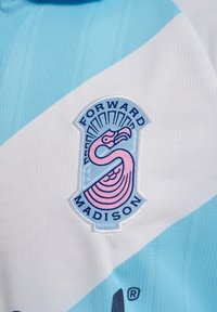 Ljust blått och vitt mönstrat tyg med en broderad logotyp med en rosa fågel symbol, omgiven av mörkare färger, på en sporttröja.