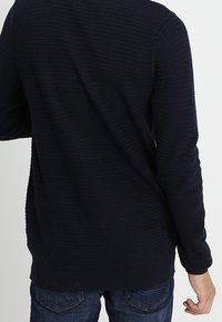 Marinblå stickad tröja med en strukturerad mönster, rund halsringning och ribbad fåll. Kombinerad med mörkblå jeans, vilket ger en avslappnad stil.