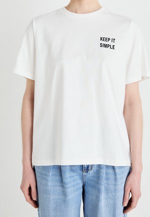 Personne portant un T-shirt blanc avec le texte "KEEP IT SIMPLE" sur la poitrine gauche, associé à un jean taille haute bleu clair.