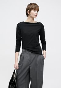 Μαύρο τοπ με μακριά μανίκια και λαιμόκοψη καραβόπανο, συνδυασμένο με γκρι πλισέ παντελόνι. Το μοντέλο κρατάει μια μαύρη γυαλιστερή τσάντα, παρουσιάζοντας μια μινιμαλιστική εμφάνιση.