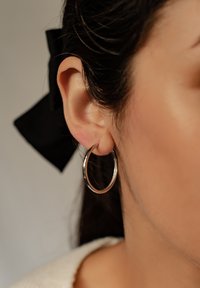 Pendiente de aro de plata con un acabado pulido, que presenta una forma circular. El fondo incluye una cinta negra y una textura de tela suave.