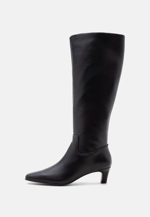 Botte noire en cuir jusqu'au genou avec un bout pointu et un petit talon incurvé sur fond blanc.