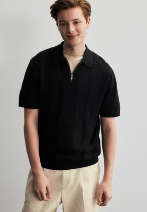 Short-sleeve black polo shirt with a zippered collar, featuring ribbed texture and vertical stripes. Worn over a light beige shirt.