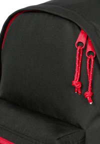 Mochila negra con parte superior redondeada, que presenta tiradores de cremallera rojos y acentos de cordón. Tela duradera con una textura suave.