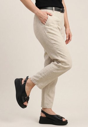 Person, die beige knöchellange Hose, schwarzen Gürtel und schwarze Plateausandalen mit Schnallendetail trägt, steht auf einem Bein vor einfarbigem Hintergrund.