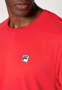 Punane spordipepup, sile tekstuur, ümmargune kaelus, millel on väike bränditud logo vasakul rinnal ja raglanvarrukad.
