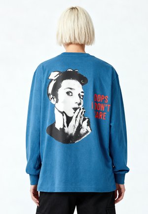 Personne aux cheveux blonds courts portant un sweat-shirt bleu avec un motif rétro en noir et blanc représentant une femme et le texte rouge « OOPS I DON’T CARE » dans le dos.