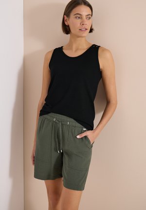 Femme portant un haut noir sans manches et un short vert texturé à cordon, debout avec une main dans la poche devant un mur beige.