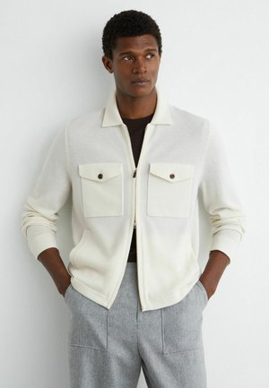 Muž v bielej bunde na zips s gombíkmi na náprsnom vrecku a sivých nohaviciach, stojaci s rukami vo vreckách, pozerajúci sa bokom.