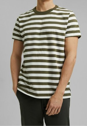 Homme portant un t-shirt à manches courtes rayé horizontalement vert olive et blanc avec un pantalon foncé sur un fond gris uni.
