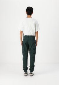 Grønne cargo bukser med sidelommer, avtagende ben og elastiske mansjetter, kombinert med en lys grå t-skjorte. Minimalistisk design, ingen synlige mønstre.