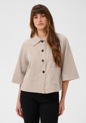 Femme aux longs cheveux bruns porte une veste beige boutonnée à manches larges et un pantalon noir sur un fond uni.