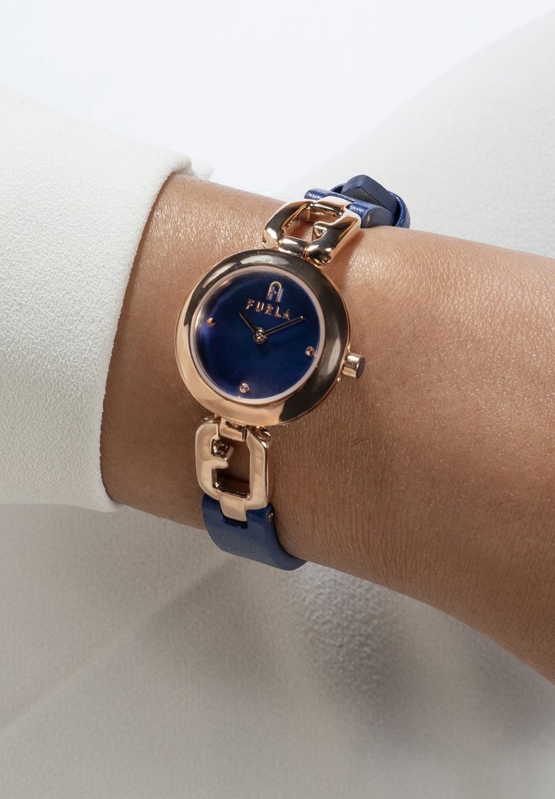 Montre-bracelet avec un boîtier rond en or rose, un cadran bleu et un bracelet en cuir bleu portée au poignet d'une personne avec une manche blanche.