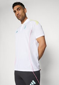 Hvid polo-shirt med grønne og gule striber på skuldrene, lyserød kant og et diskret logo på brystet; parret med mørke bukser.