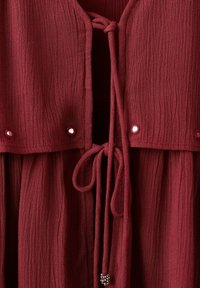 Close-up van een bordeauxrode jurk van gestructureerde stof met voorstrikken en kleine metalen kraaltjes aan het uiteinde en langs het borstgebied.