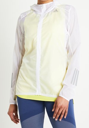 Persoon draagt een wit en bleekgeel Adidas-jas met ritssluiting met reflecterende strepen op de mouwen en blauwe legging met patroon.