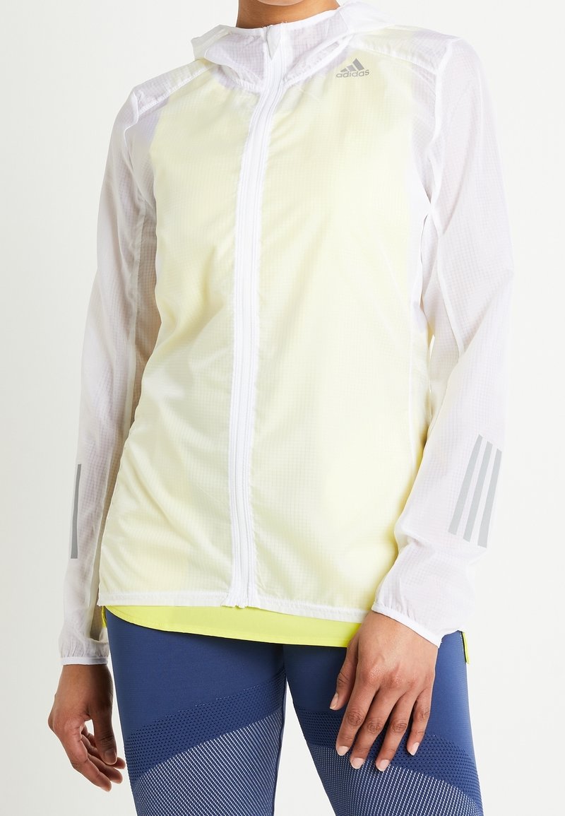 Personne portant une veste zippée Adidas blanche et jaune pâle avec des bandes réfléchissantes sur les manches et des leggings bleus à motifs.