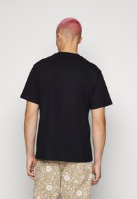 T-shirt nera in cotone con maniche corte, colletto a giro, e una vestibilità comoda, vista da dietro, abbinata a pantaloni corti beige con motivo.
