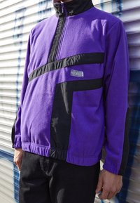 Lilla fleecejakke med sorte paneler, halv-zip krave, elastiske manchetter og et synligt logo på brystet. Struktureret materiale.