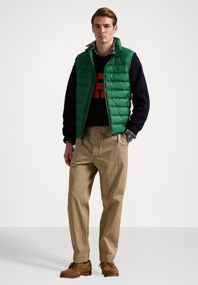 Green quilted vest over a dark sweater with a large orange emblem, beige trousers, and brown moccasin-style shoes.