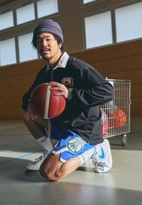 Pantalones cortos deportivos en azul con un logotipo de baloncesto verde, camiseta de manga larga negra y zapatillas Nike blancas, sosteniendo un balón de baloncesto rojo.