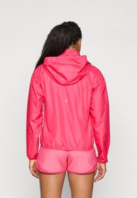 Letvægts pink vindjakke med hætte, elastiske manchetter og søm. Udstyret med et lille logo på ryggen. Kombineret med pink shorts.