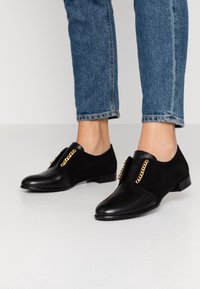 Chaussures noires en cuir lisse et en daim, avec des accents en chaîne dorée sur le dessus et un petit talon. Portées avec un jean bleu.
