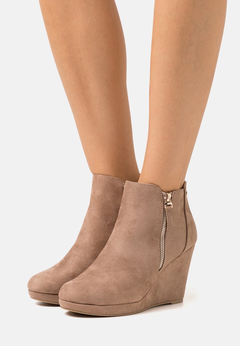 Bottines beige en daim à talon compensé avec fermeture éclair latérale, portées sur jambes nues sur un fond uni.