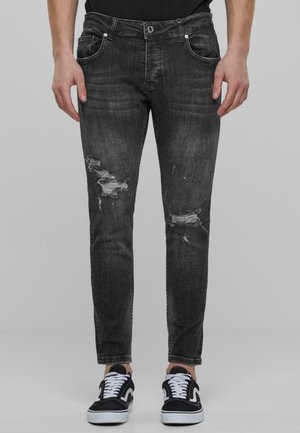 Jeans ajustados en denim gris oscuro con detalles desgastados. Cuenta con cinco bolsillos y un cierre de botón. Combinados con zapatillas negras y blancas.