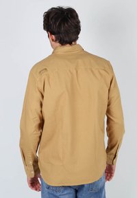 Chemise beige à manches longues, au design simple, avec un petit logo dans le dos. Le tissu semble doux et léger.