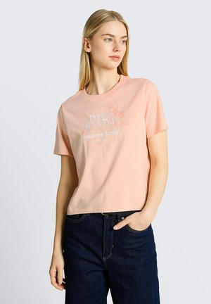 Junge Frau mit langen blonden Haaren, die ein hellrosa bedrucktes T-Shirt und dunkelblaue Jeans trägt, schaut zur Seite mit einer Hand in der Tasche.