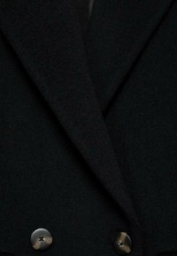 Gros plan des revers d'un manteau noir avec deux grands boutons foncés, mettant en valeur la texture et les détails des boutons.