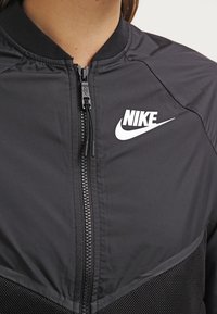Zwarte Nike-jas met een ritssluiting aan de voorkant, voorzien van een witte logo en een gestructureerd mesh paneel aan de onderkant. Gladde stof met een geribde kraag.
