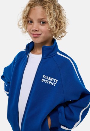 Veste zippée bleue avec des empiècements blancs et le texte brodé "YOSEMITE DISTRICT" sur le côté gauche. Tissu doux, coupe décontractée, col élevé.