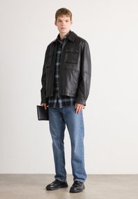 Black leather jacket with two chest pockets, blue plaid shirt underneath, light blue jeans, and black shoes. Model holding a black clutch.