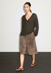 Una donna cammina con sicurezza, indossando un maglione di maglia scuro, una gonna di suede marrone e sandali, su uno sfondo minimalista.