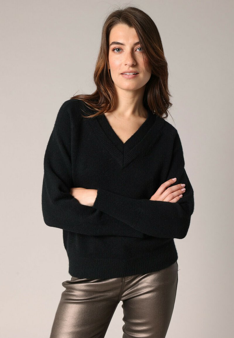 Femme aux cheveux bruns portant un pull noir en V et un pantalon bronze métallisé, se tenant les bras croisés contre un fond uni.