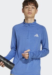 Modrá športová košeľa s dlhým rukávom, vysokým golierom a štvrtinovým zipsom. Má otvory na palce v rukávoch a biele logo Adidas na hrudi.