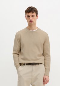 Jeune homme portant un pull côtelé beige sur une chemise blanche et un pantalon beige avec une ceinture foncée, debout devant un fond blanc uni.