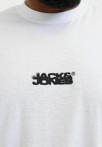 Bijela pamučna majica s crnim logotipom "JACK & JONES" u stilu spreja na prednjoj strani. Okrugli izrez i mekana tekstura.